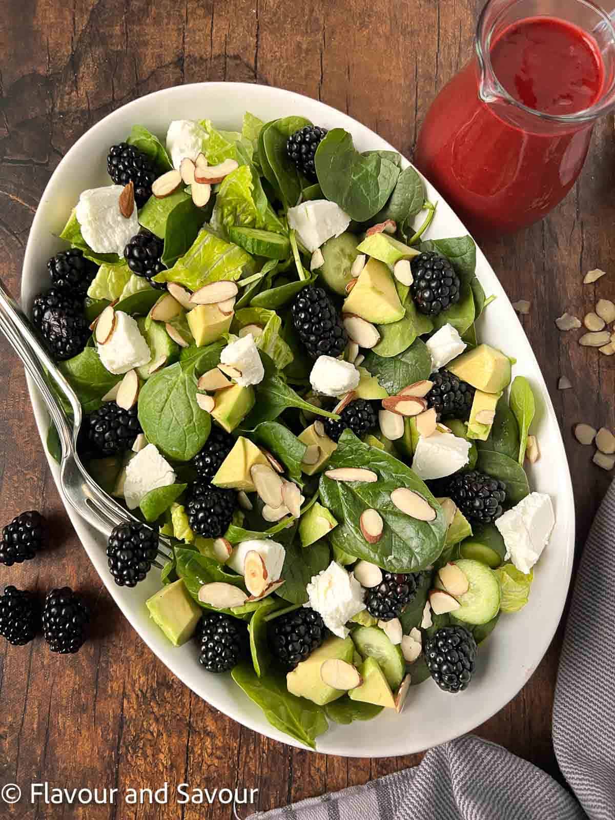 Spinach salad with blackberries, goat cheese, avocado and toasted walnuts in a bowl with a small pitcher of blackberry dressing beside.