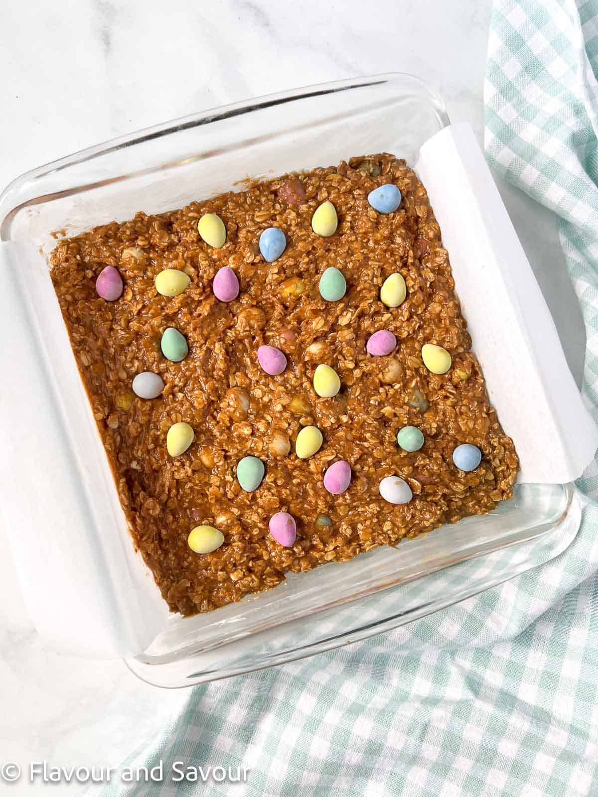 Mini egg cookie bar dough spread in a baking pan with mini eggs sprinkled randomly on top.