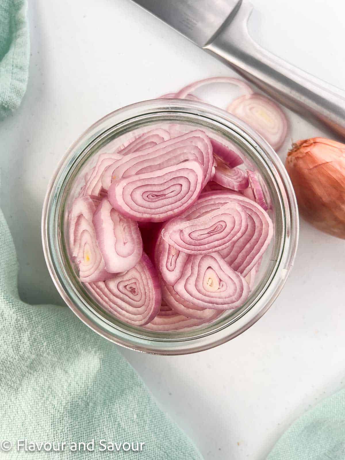 Sliced shallots in a jar with a knife and a shallot beside the jar.