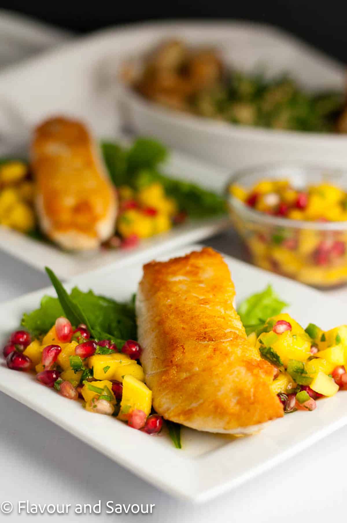 Two plates with salt and pepper crusted halibut fillets served with mango-pomegranate salsa in a small bowl beside.