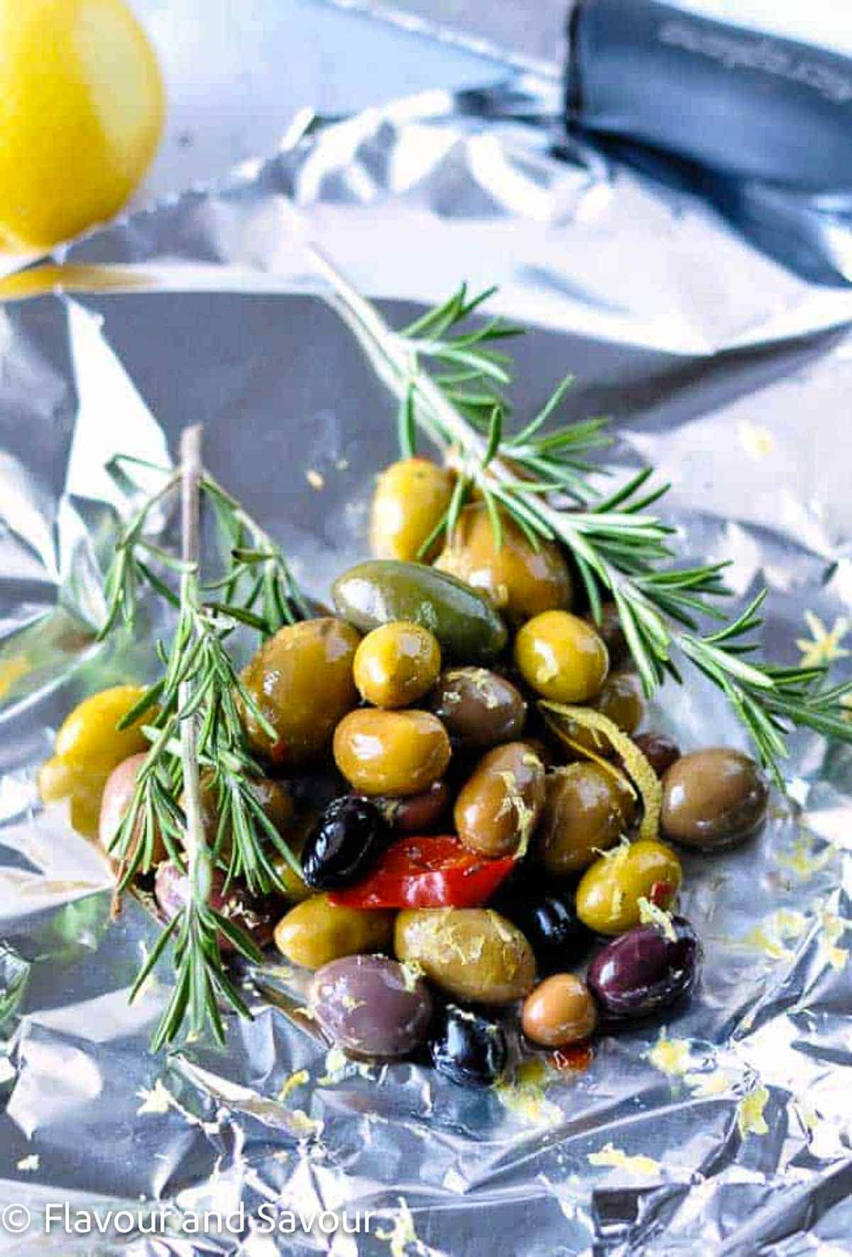 A small pile of mixed olives on a sheet of aluminum foil with fresh rosemary sprigs.