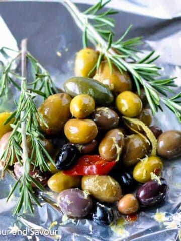 Mixed olives in small pile on a sheet of aluminum foil with sprgis of rosemary.